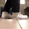 A close-up view of a person wearing white safety boots and dark pants, standing on a tiled floor. Produktbild-Vorschau