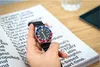 A hand holding a watch over a book with text. Produktbild-Vorschau