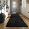 A black rug is laid out on a wooden floor in a hallway, with a large black vase and a plant on the left. Produktbild-Vorschau