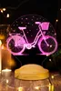 A pink 3D LED lamp shaped like a bicycle with a basket, displayed on a wooden base with decorative lights and candles in the background. Produktbild-Vorschau
