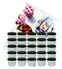 A set of small glass jars with black lids, along with ingredients for making jam, including strawberries, a packet of gelling sugar, and a checkered cloth. Podgląd zdjęcia produktu
