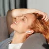 A woman with red hair is lying down with her eyes closed and a smile on her face, wearing white wireless earbuds. Náhled obrázku produktu