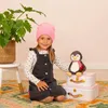 A young girl sits on a rug next to a stuffed penguin and two decorative suitcases, with a large potted plant in the background. Anteprima dell’immagine prodotto