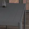 A close-up view of a dark gray outdoor dining table with a glass vase containing decorative items on its surface. The table is set on a wooden deck, with a blurred background of a window and wooden flooring. Produktbild-Vorschau