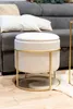 A beige ottoman with a gold metal frame sits on a wooden floor in front of a gray sofa with pillows. Náhled obrázku produktu