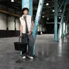 A person stands in a large, industrial-looking space, holding a black basket. Voir les images du produit