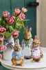 Three ceramic bunnies are displayed on a white surface in front of a bouquet of pink tulips and green foliage, with a green wooden door in the background. Podgląd zdjęcia produktu