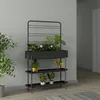 A black metal plant stand with a planter box and shelves, holding several potted plants and flowers, placed against a light gray wall. Podgląd zdjęcia produktu