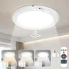 A white LED ceiling light with a motion sensor and remote control is shown mounted on a white ceiling. The light is emitting a beam downwards. Below the main image, there are three smaller inset images showing the light with different color temperatures. T Produktbild-Vorschau