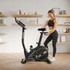 A woman is adjusting the seat of an exercise bike in a room with shelves filled with books and decorative items. Podgląd zdjęcia produktu