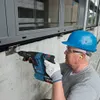 A construction worker is using a Bosch cordless hammer drill to drill into a concrete wall next to a window. Produktbild-Vorschau