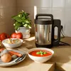 A kitchen counter with a soup maker, ingredients, and a bowl of soup. Produktbild-Vorschau
