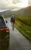 A person rides a bicycle on a road next to a car, with mountains in the background. Náhled obrázku produktu
