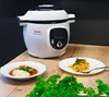 A Tefal Cook4me+ smart multi-cooker is shown on a wooden table, with two plates of food in front of it and some greenery in the foreground. Podgląd zdjęcia produktu