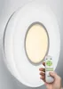 A hand holding a remote control is shown next to a white, circular LED ceiling light mounted on a wall. Produktbild-Vorschau