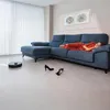 A woman is sleeping on a blue sectional sofa while a robot vacuum cleaner cleans the floor in the foreground. A pair of black high heels are also on the floor. Náhled obrázku produktu