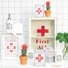 A collection of decorative items with a medical or pharmacy theme, including a medicine cabinet, soap dispenser, toilet paper, and small potted plants, arranged on a white surface against a tiled background. Produktbild-Vorschau