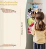 A young girl looks at a hanging shelf filled with stuffed animals, with a ruler indicating the height. Produktbild-Vorschau