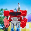 A toy Optimus Prime robot with a small human figure standing on its head, set against a blurred outdoor background of trees and houses. Podgląd zdjęcia produktu