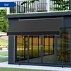 A black awning is extended over a glass sliding door on a building, with a balcony above and a garden in the foreground. Náhľad obrázku produktu