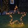 A star-shaped garden light decoration with warm white LEDs is placed on a lawn in front of a dark-colored building with a lit window. Produktbild-Vorschau