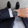 A person wearing a suit and watch, with their shoes visible. Produktbild-Vorschau