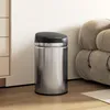 A silver and black sensor trash can is placed on a wooden floor next to a beige sofa and a cabinet. Produktbild-Vorschau