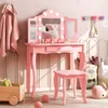 A pink children's vanity table and stool set with a tri-fold mirror and lights, set in a child's room with decorative pillows and a hooded towel. Produktbild-Vorschau