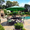 An outdoor patio scene with a table and chairs set up under a large green umbrella, next to a swimming pool and a stone fireplace. Produktbild-Vorschau