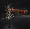 A decorative LED tree with white lights stands in a snowy yard in front of a red building at night. Podgląd zdjęcia produktu