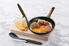 A frying pan with food and utensils on a wooden board, set against a marble background. Voir les images du produit