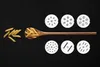 A wooden spoon filled with penne pasta is placed on a black background, next to a pile of uncooked penne pasta. Six white discs with various hole patterns are arranged above and below the spoon. Produktbild-Vorschau