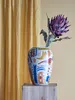 A decorative vase with an abstract pattern and a single flower is placed on a wooden stand in front of a yellow curtain. Náhled obrázku produktu