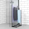 A black metal towel rack with three towels hanging on it, placed in a modern bathroom with white subway tiles and gray floor tiles. Náhľad obrázku produktu