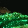 Green net lights are draped over bushes at night, with a partial view of an umbrella in the background. Voir les images du produit