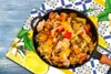 A close-up of a cooked vegetable dish in a pan, placed on a decorative cloth with a lemon motif. Produktbild-Vorschau