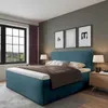 A bedroom scene featuring a blue upholstered bed with a brown throw blanket and white bedding. There is a window with purple curtains on the left and a nightstand with a lamp on the right. Three framed pictures hang on the wall above the bed. Produktbild-Vorschau