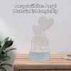 A 3D acrylic night light shaped like a teddy bear holding a heart-shaped balloon sits on a wooden table next to a plant. The background is a plain wall. Produktbild-Vorschau