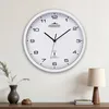 A white analog wall clock with black numbers and hands is mounted on a white wall above a wooden shelf. Anteprima dell’immagine prodotto