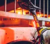 A close-up shot of a bicycle seat post with a red taillight attached, set against a blurred background of warm lights and metal railings. Podgląd zdjęcia produktu