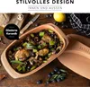 A rustic table setting with a clay pot filled with a cooked meal, surrounded by ingredients like blueberries and mushrooms. Produktbild-Vorschau