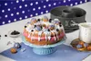 A decorated cake with pink frosting and white icing, surrounded by blueberries, small pastries, and baking equipment. Produktbild-Vorschau