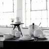A collection of white porcelain tableware, including teacups, a teapot, bowls, and a sugar bowl, arranged on a dark tablecloth in front of large windows. Produktbild-Vorschau