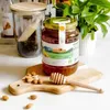 A jar of honey sits on a wooden cutting board with almonds and a honey dipper, with a glass jar and a plant in the background. Produktbild-Vorschau
