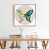 A framed embroidery artwork of a butterfly with flowers, displayed on a wall above a wooden dining table. Produktbild-Vorschau