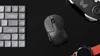 A black wireless mouse is shown on a black surface, next to a gray numeric keypad and a portion of a smartwatch with a red strap. Produktbild-Vorschau