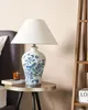 A table lamp with a blue and white porcelain base and a white conical lampshade sits on a small side table next to a bed. A wooden ladder shelf is visible on the right. Náhľad obrázku produktu
