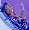 A young woman with purple and blue hair, wearing a shiny blue jacket, is lying down and holding a small pink bottle with glitter inside. Náhled obrázku produktu