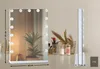 A makeup mirror with lights is displayed on a table with some makeup brushes and decorative items in the background. Podgląd zdjęcia produktu