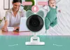 A woman is working on a laptop while a baby lies on a bed, with a baby monitor camera between them. Náhľad obrázku produktu